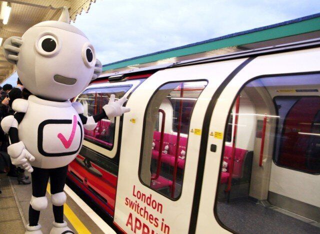 ID: A mascot costume of robot character Digit Al stands to the left of a tube train that advertises when London switches to digital television.